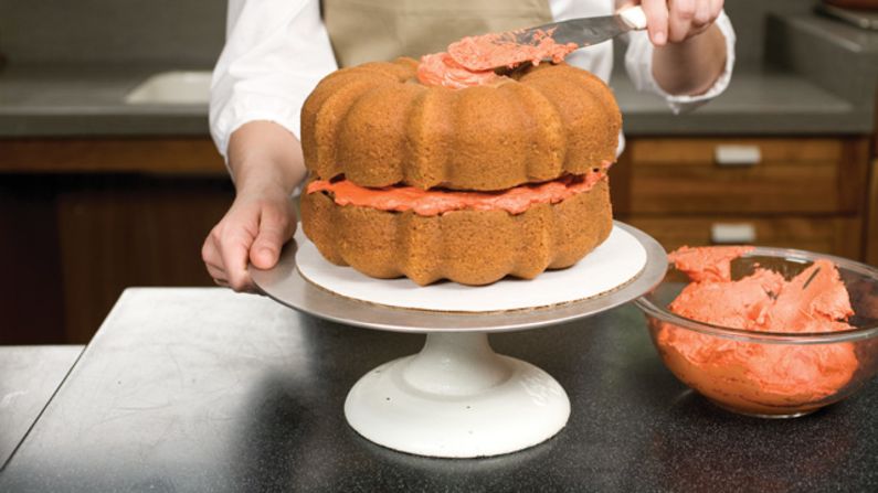 After placing one Bundt cake flat side up on a cake platter and covering the flat side with 1 cup orange frosting, place the second Bundt cake flat side down on top of the frosting, making sure the ridges on both cakes align. Then place one cupcake cut side up in the hole of the Bundt cake and spread 1 tablespoon orange frosting over the top of the cupcake. <a href="http://eatocracy.cnn.com/2013/10/24/pumpkin-cake/">See the full recipe.</a>