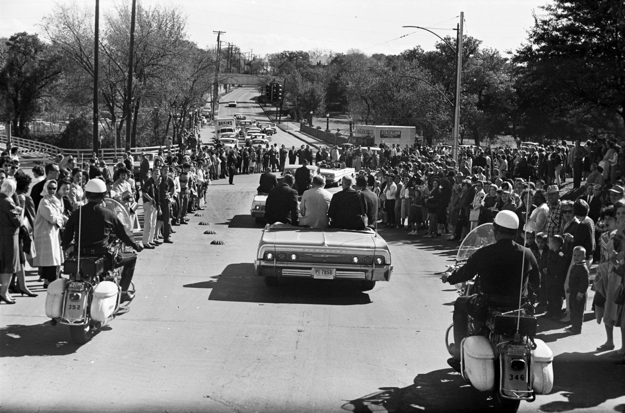 Crowds line the street as Kennedy's motorcade heads toward downtown Dallas. A group of White House staffers follows the motorcade in a bus several vehicles behind the presidential limousine. 