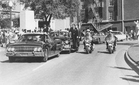 Seen through the limousine's windshield as it proceeds along Elm Street past the Texas School Book Depository, Kennedy appears to raise his hand toward his head after being shot. The first lady holds Kennedy's forearm in an effort to aid him. 
