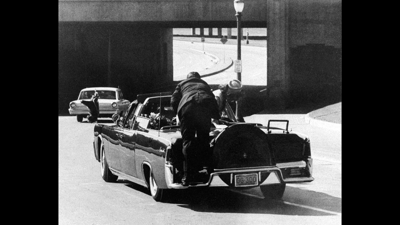Kennedy slumps in the back seat of the car and his wife leans over to him as Secret Service Agent Clinton Hill rides on the back of the car.