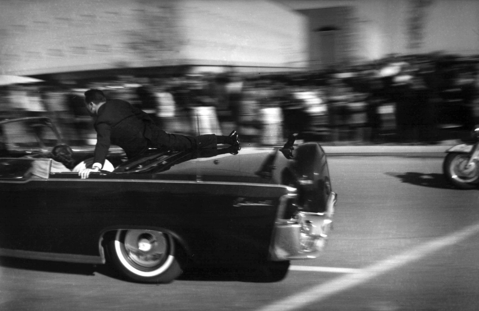 The limousine carrying the mortally wounded president races toward the hospital seconds after the shots were fired. Here, Secret Service agent Clint Hill rides on the back of the car as the wives cover their stricken husbands.