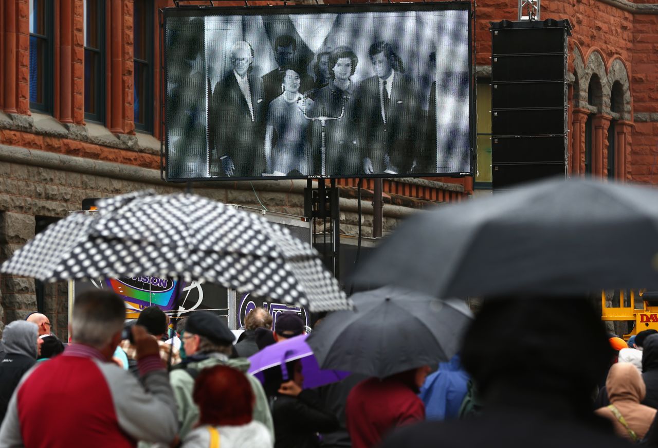 People near Dealey Plaza watch a historical broadcast about Kennedy's life.