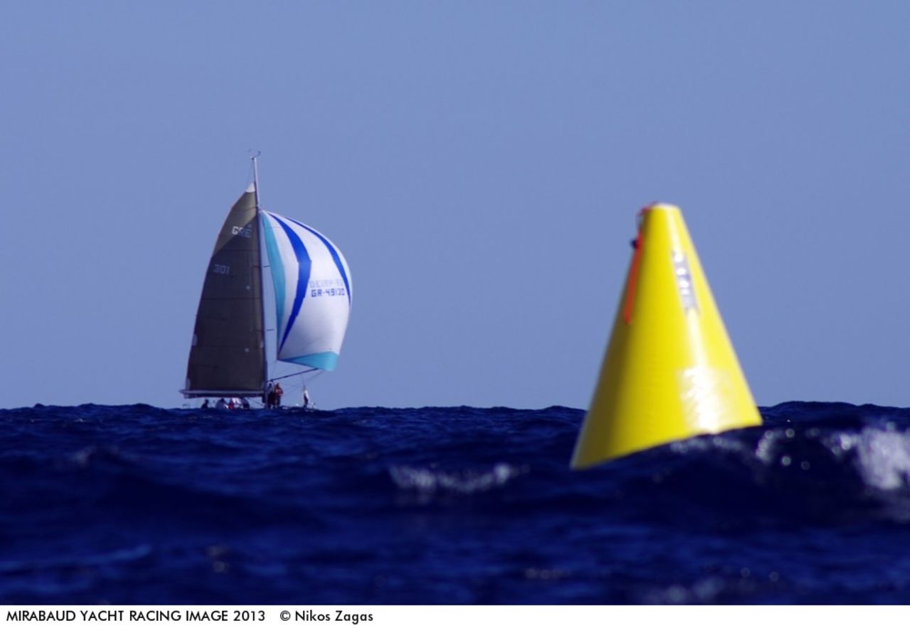 An out-of-focus yellow buoy bobs on the surface of the water as, in a distance, Pachakis Ioannis glides over the water in October's Cretan Union Cup, captured on a Pentax 450mm.