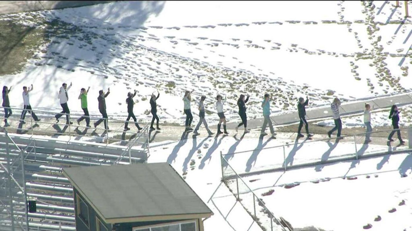 Students walk away from the school in single file with their hands up.