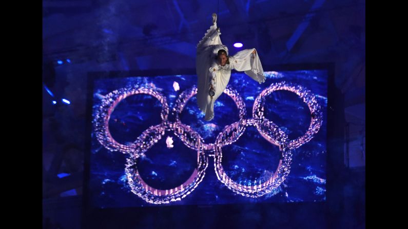 A performer hangs from a wire in front of the Olympic rings.