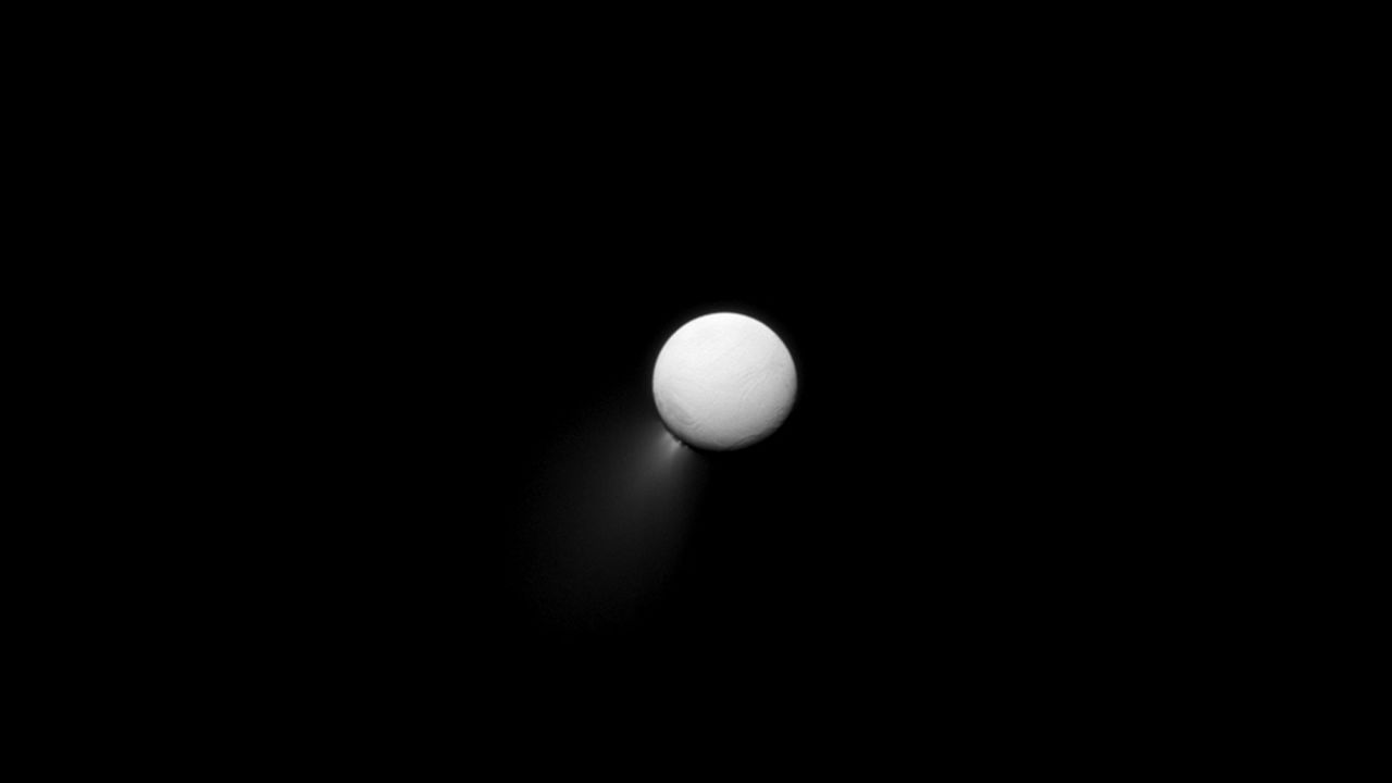 The Saturn-facing side of Enceladus is illuminated by light bouncing off the planet. Plumes of water ice can be seen streaming off the moon's southern pole.