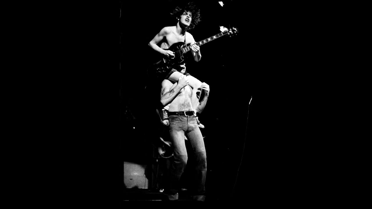 Angus Young sits on Bon Scott's shoulders while performing at the Town Hall in St. Albans, England, in 1976. Young is known for performing in a schoolboy uniform.