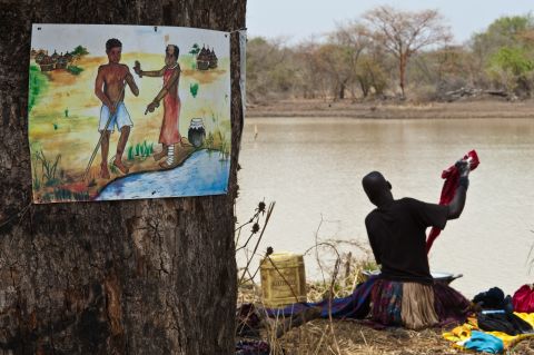 In South Sudan, a woman washes clothes in a roadside dam next to a sign warning of the dangers of spreading Guinea worm disease by entering the water to ease the burning pain caused by an emerging worm. 
