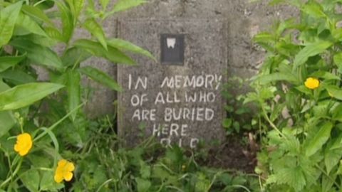 A marker to those buried at a former home for unwed mothers sits against a stone wall. 