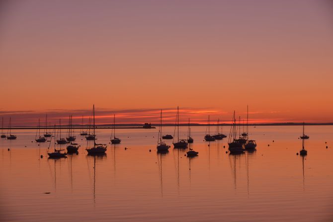 Yachts and sailboats sit anchored at sunrise in Maine's Rockland Harbor with the Rockland Breakwater Lighthouse in the background. The harbor offers ferries to Vinalhaven and other islands.