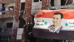 Caption:THIS PICTURE WAS TAKEN ON A GOVERNMENT-GUIDED TOUR A flag showing Syrian President Bashar al-Assad flutters near damaged buildings in Adra northeast of the capital Damascus on September 25, 2014. Syrian government troops recaptured an area of the key rebel-held town Adra used to house workers, after securing the highway and the industrial zone, a security source and monitoring group said. AFP PHOTO / LOUAI BESHARA (Photo credit should read LOUAI BESHARA/AFP/Getty Images)