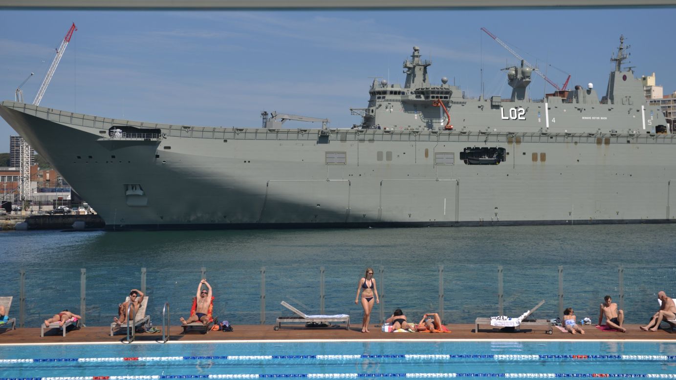 People in Sydney sunbathe at a swimming pool in front of HMAS Canberra, the Royal Australian Navy's latest warship, on Friday, November 21.