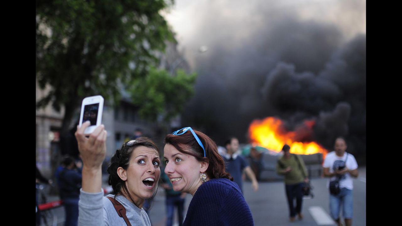 Tourists take a selfie as demonstrators burn a trash container during a May Day rally in Barcelona, Spain, on Thursday, May 1. May Day is also known as International Workers' Day, and tens of thousands of workers in European cities marked the occasion with a mix of anger and gloom over austerity measures meant to contain the eurozone debt crisis.