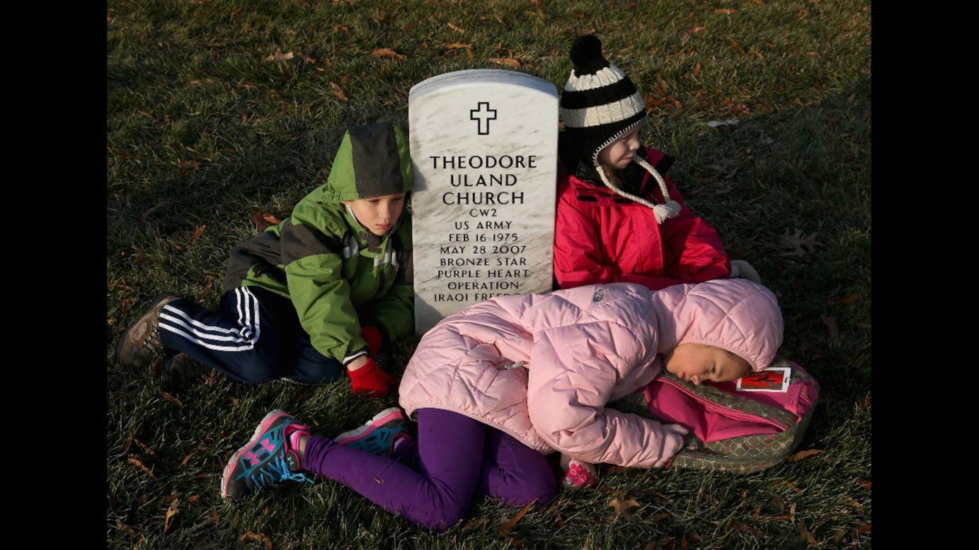 From left, Jack Harris, Jada Harris and Olivia Bernhardy sit at the grave of a close family friend Saturday, December 13, at Arlington National Cemetery in Arlington, Virginia.