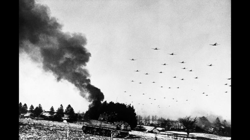 An American tank burns while transport planes fly overhead during the Battle of the Bulge.