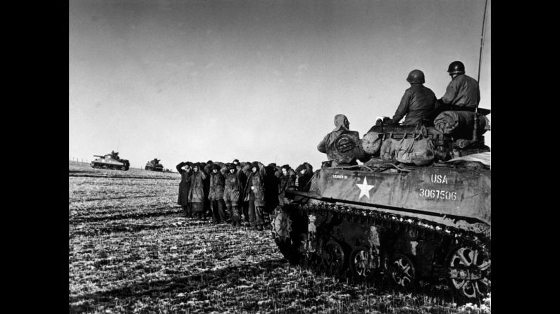 American troops ride on a tank while German POWs are held nearby.