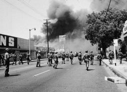 Armed National Guardsmen march toward smoke on the horizon during the street fires of the Watts riots, Los Angeles, August 1965. 