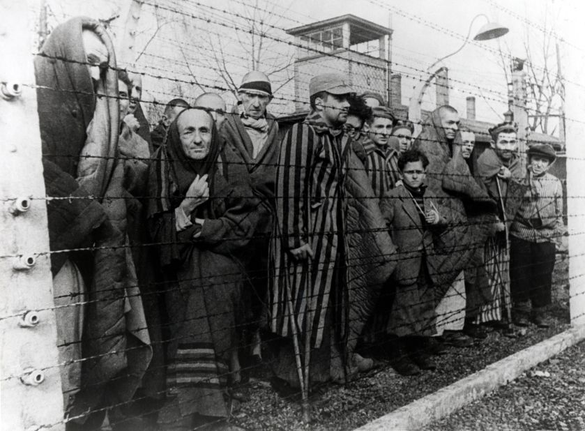 Survivors of Auschwitz stand behind a barbed wire fence. Some of the children are seen wearing adult clothing they were dressed in by Soviet soldiers. 