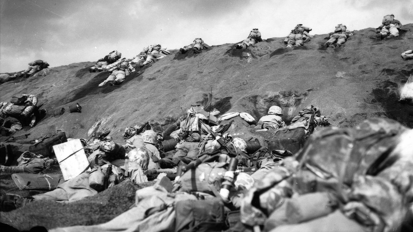 In the face of enemy fire, Marines inch their way over the slope on Red Beach No. 1. In the foreground, other Marines huddle in shell holes as they wait for orders to move forward.