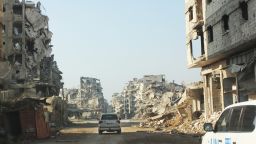 On 24 January 2015 in the Syrian Arab Republic, vehicles of the United Nations travel a dusty road lined with the rubble remnants of destroyed buildings, in the Old City of Homs, Homs Governorate. One of the vehicles bears the UNICEF logo.
