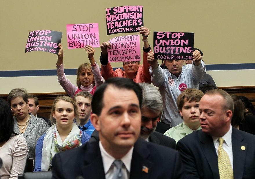 Members of Code Pink (left to right) Medea Benjamin, Liz Hourican and Tighe Barry, hold signs to protest as Walker (center) takes his seat during a hearing before the House Oversight and Government Reform Committee April 14, 2011, on Capitol Hill in Washington, D.C.