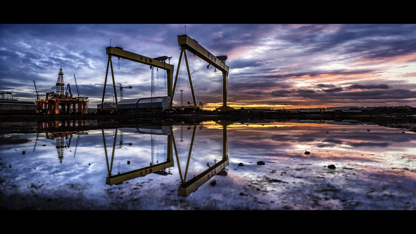 <strong>"Perfect Symmetry" by Norman Quinn</strong><br />Photographer's description: The two great yellow-painted gantry cranes Samson and Goliath have become icons of Belfast (Northern Ireland), dominating not just Queen's Island but the entire city skyline. Constructed to service the vast new graving dock at Harland and Wolff, Goliath (the smaller at 315 feet) began work in 1969; the 348-foot Samson, five years later. Each of the Krupp-Ardelt-designed cranes can lift loads of up to 840 tons. Harland and Wolff were still one of the world's great shipbuilders at the time, and the building of the two cranes, during a difficult period for shipbuilding in Belfast, was seen as a sign of faith in the future. The image itself was taken during sunrise on a particularly wet day by lying flat in a puddle as low as possible to the ground. It is a series of images stitched together in editing software to create an impressive colorful panoramic.