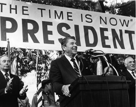 Reagan smiles as he speaks about his presidential campaign in 1979 front of a large banner with his campaign slogan, "The Time is Now."
