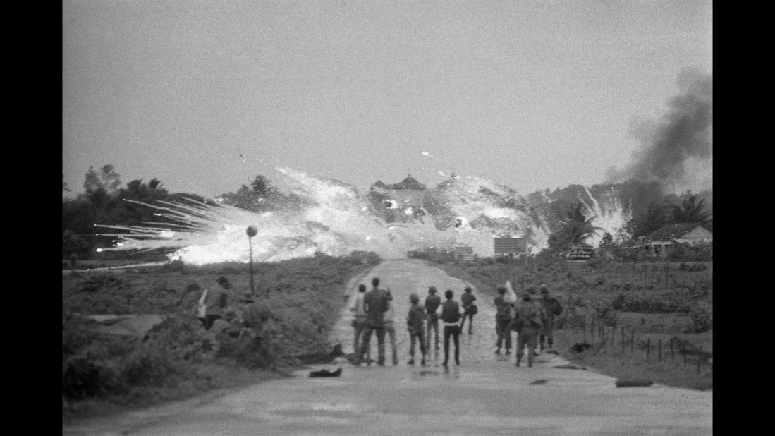 As bombs drop in Trang Bang, soldiers and members of the international media watch the scene in the foreground.