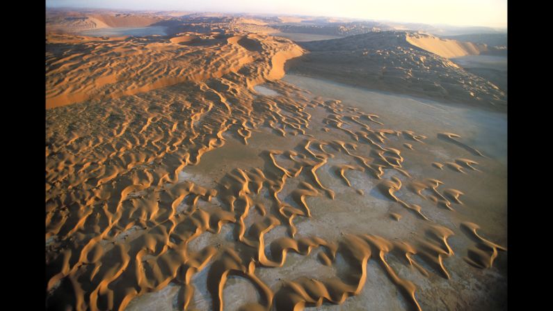 A salt flat in Saudi Arabia has crescent-shaped barchan dunes marching across its basin in 2002. The orange color of the dunes is due to oxidation of iron minerals in the sand.