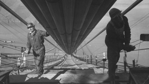 Two workmen add the last strands to one of the two enormous cables that run between the towers and support the six-lane highway below. Construction of the bridge cost $35 million; engineers estimate that if it was built today, the bridge would cost more than $1.3 billion.