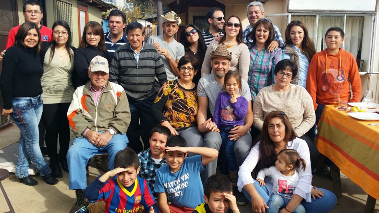 Travis Tolliver sits in the center surrounded by his biological family in Chile. He's wearing a hat and a young girl in a purple T-shirt sits on his lap. 