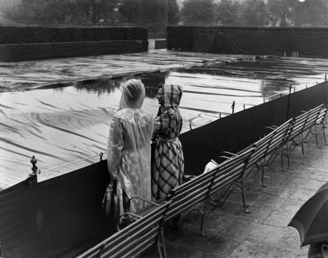 Two spectators gaze forlornly at flooded, covered Wimbledon courts in July 1950.