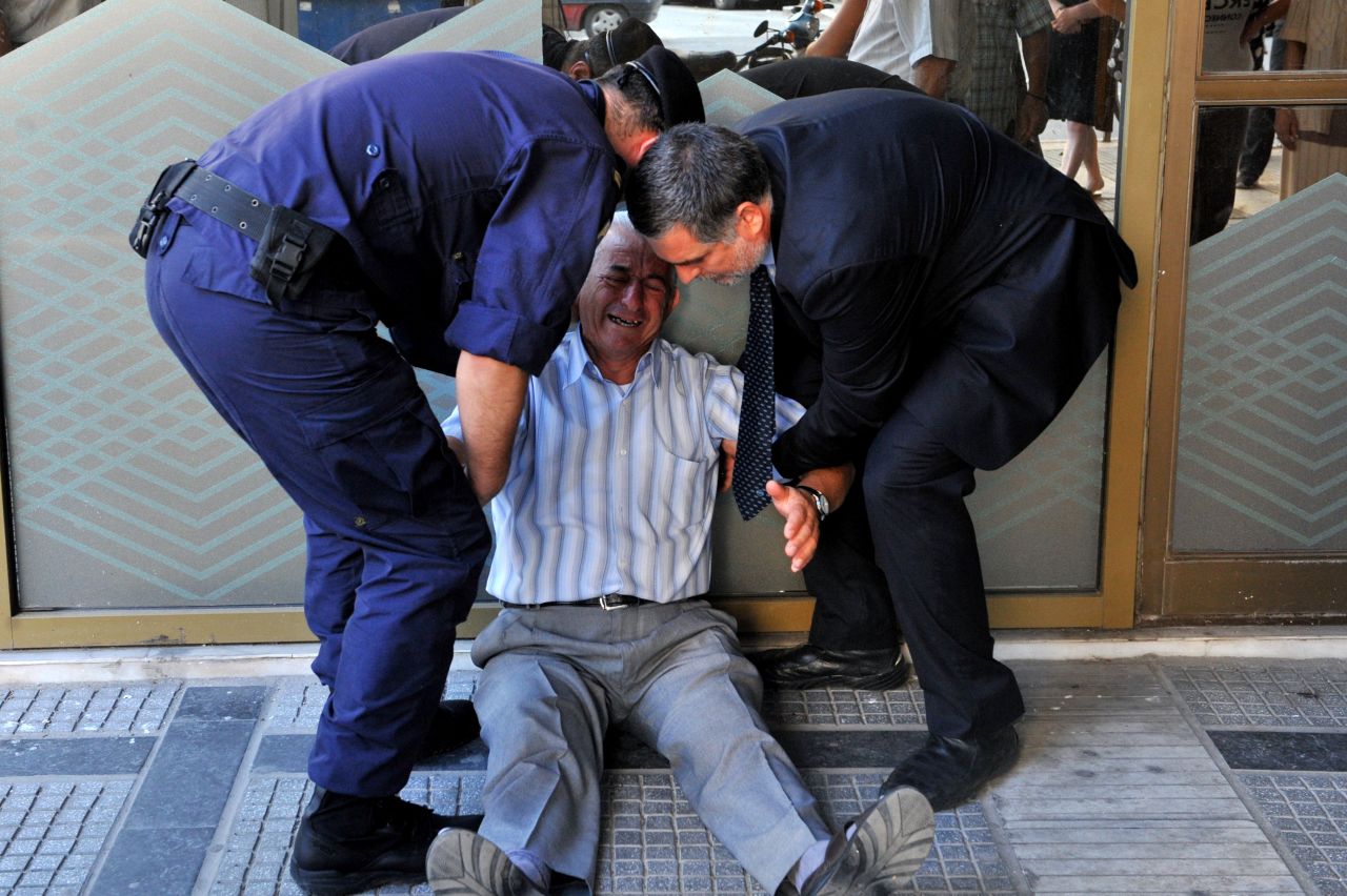 An crying elderly man is assisted by an employee and a policeman outside a national bank branch as pensioners queue to get their pensions, with a limit of 120 euros.