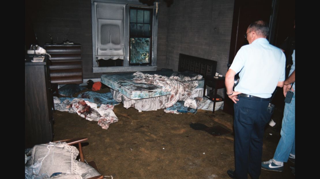 A crime scene photo shows two police officers inspecting the burnt-out bedroom where Karen and Dyke Rhoads had been murdered.