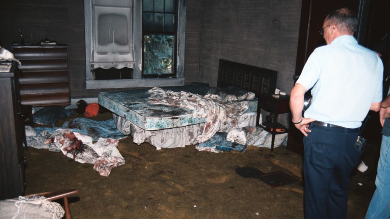 A crime scene photo shows two police officers inspecting the burnt-out bedroom where Karen and Dyke Rhoads had been murdered.