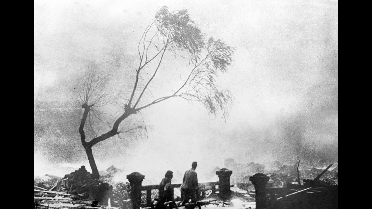 Survivors of the Nagasaki bomb walk through the destruction as fire rages in the background.