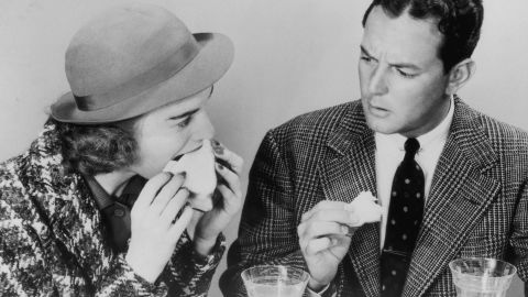 A man expresses disapproval at his friend's table manners as she sinks her teeth greedily into a sandwich, circa 1940. 