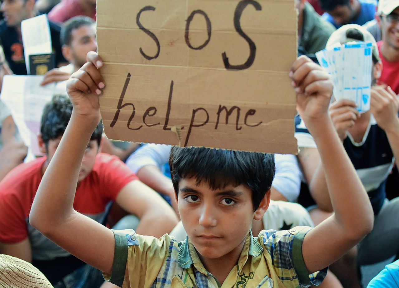A boy holds a sign reading "SOS help me" in front of Keleti station.