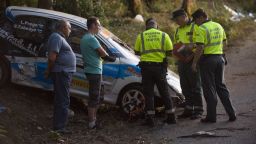 Investigators stand beside a damaged rally car near the village of Carlal.