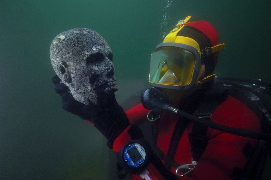 Here, a diver inspects a recovered black granite statue depicting a priest's head. The scalp is probably shaven, as was befitting of priests, for reasons of purity. The cheekbones are pronounced and the cheeks hollowed. On the forehead, the scarification mark of priests is visible and the eyes, now hollowed out, would have been incrusted with something unknown today. 