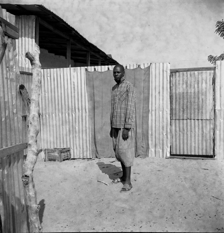 The setting and proportions mark this as a formal portrait of a man believed to be a mason in his everyday work wear, the photographer's wide lens drawing out each element of the sitter's environment.<br /><br />Man Standing in a Courtyard, 1959-1968<br />Oumar Ka (Senegalese, b. 1930)<br />Inkjet print, 2015