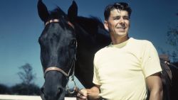A 1935 portrait of American actor Ronald Reagan in a yellow t-shirt standing next to a black horse holding its reins.