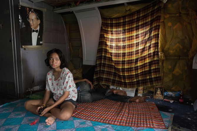 The interior of the planes occupied by the families show a few rudimentary decorations. Here a young woman sits under a picture of the Thai king, while makeshift curtains cover the windows.  