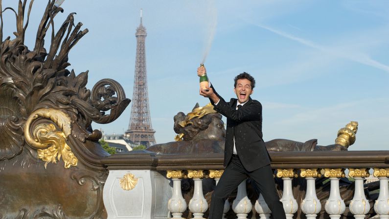 Stop with all the Paris cliches, says comedian Olivier Giraud, pictured here standing in front of the Eiffel Tower. With a bottle of Champagne.