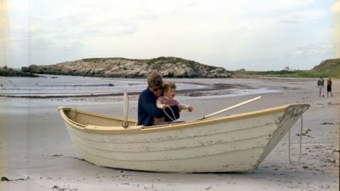 Kennedy and his son, John Jr., in a rowboat in Newport, Rhode Island. Benjamin C. Bradlee, editor of the Washington Post, and Antoinette Bradlee walk in the background.