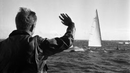 President John F. Kennedy waves to the crew of the yacht, "Weatherly" (of the New York Yacht Club), while watching the first race of the 1962 America's Cup aboard the USS Joseph P. Kennedy, Jr., off the coast of Newport, Rhode Island.