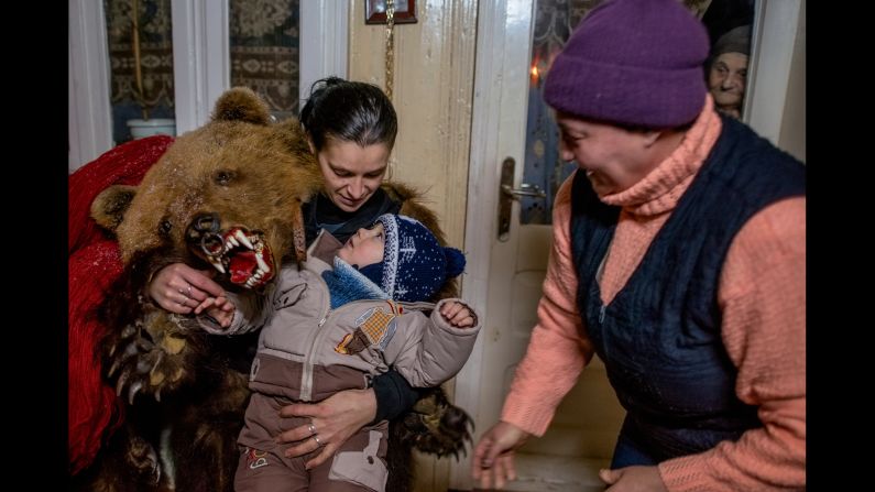 A female bear plays with the young son of Catalin Apetroaie, a bear himself. The child's grandmother scurries by while his great-grandmother peers from inside the house.  