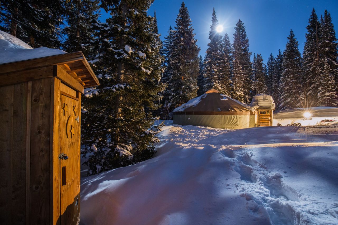 Illuminated by lanterns and moonlight, the yurt is set in a forest clearing, surrounded by tall pine trees.