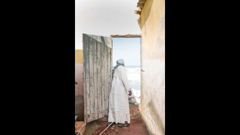 A man looks out at the sea through a door that once led to another room of his house. He and his family moved furniture out the front door, while the sea chewed at the back.