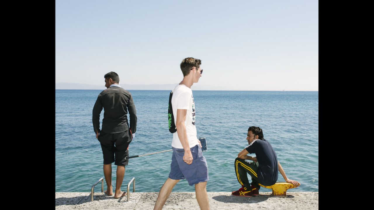 A German tourist, with selfie stick in hand, walks past migrants on the beach promenade.